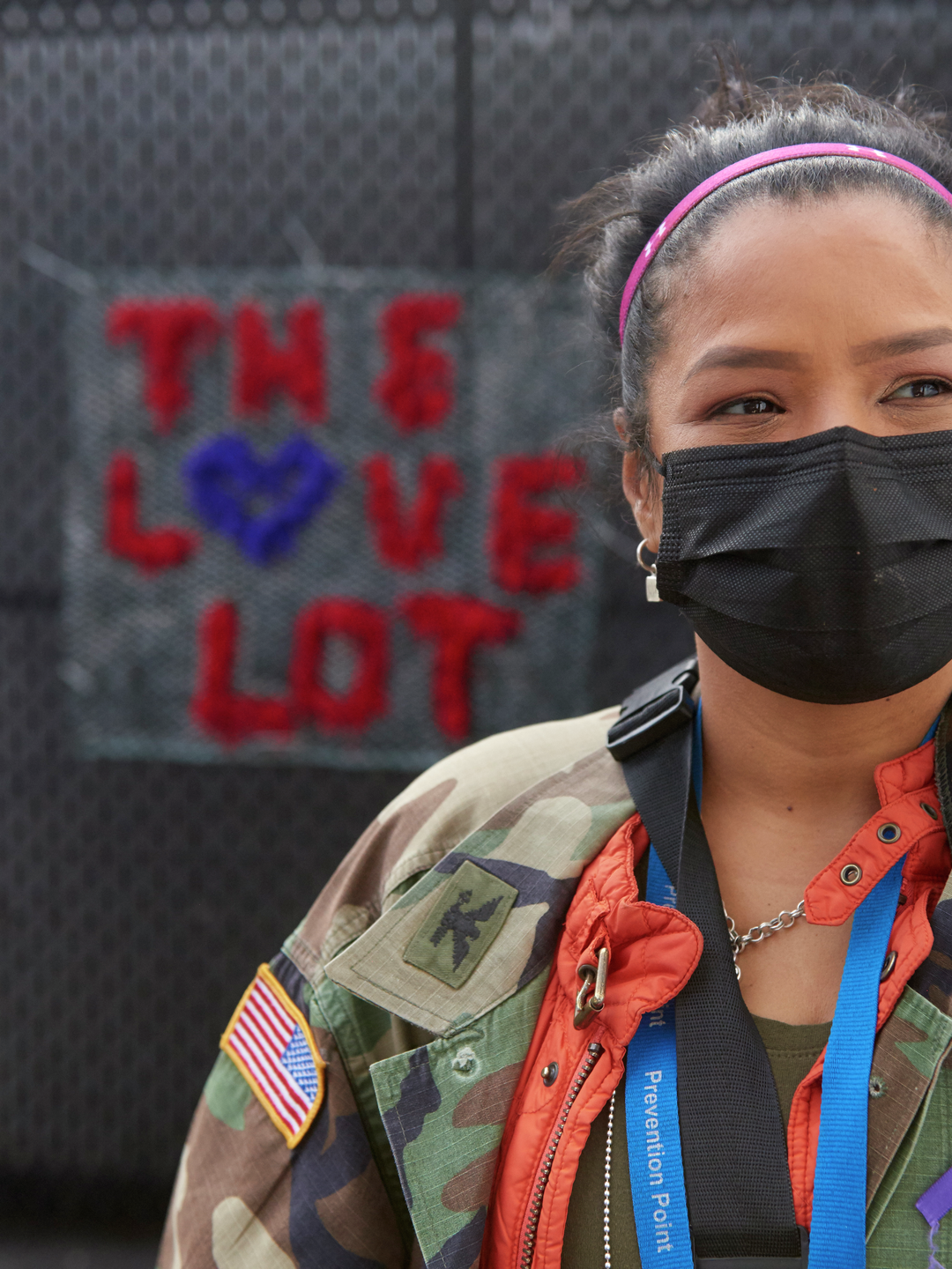 Roz Pichardo, a young woman with a face mask, and a warm but serious expression, stands in front of a crocheted sign that says "The Love Lot"