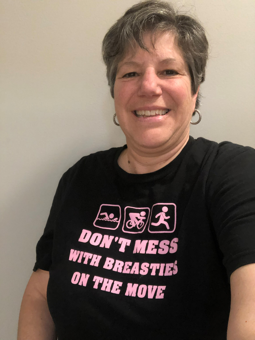Doris Piccinin poses in a black T-shirt with icons for swimming, cycling, and jogging. "Don't Mess With Breasties on the Move" is printed in pink.