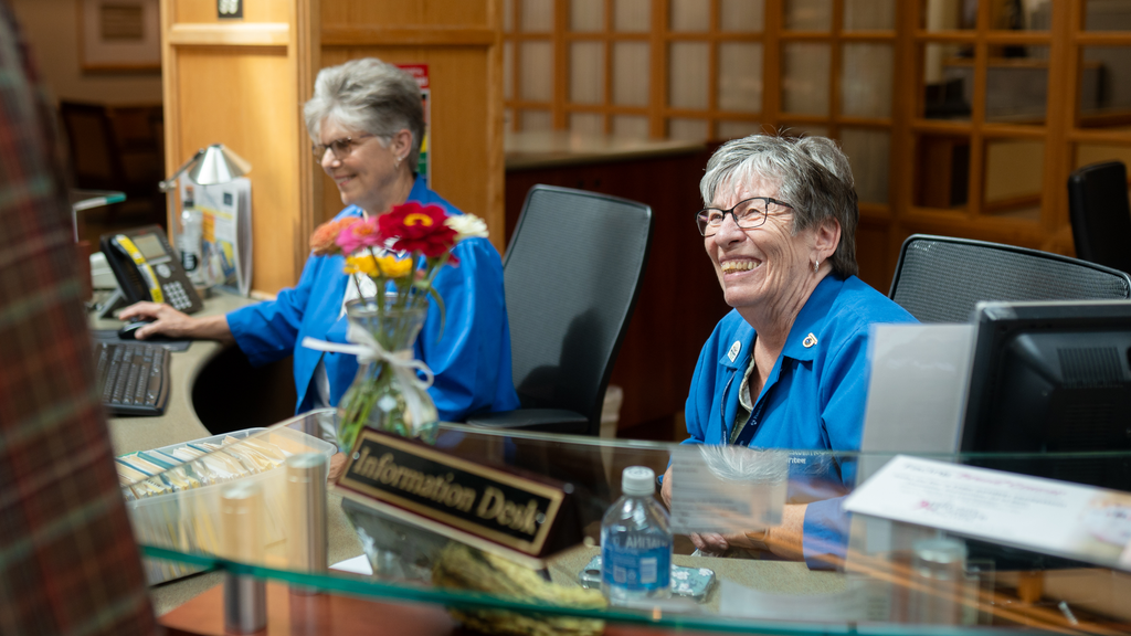 Chris Bartells and Elaine Smith, information desk volunteers, sitting at their desk and smiling while answering questions from a visitor